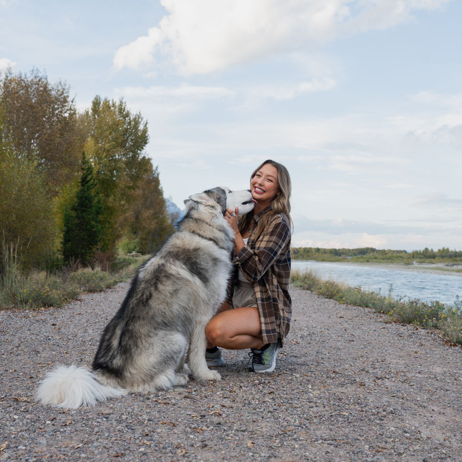 Ellie crouching beside her husky Sushi on a gravel bar along the river in fall
