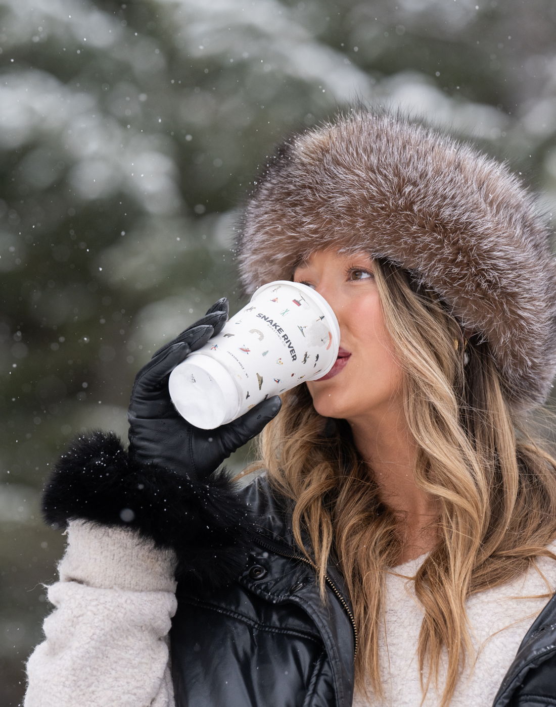 A sip of coffee from a white cup in a fur hat as snow falls