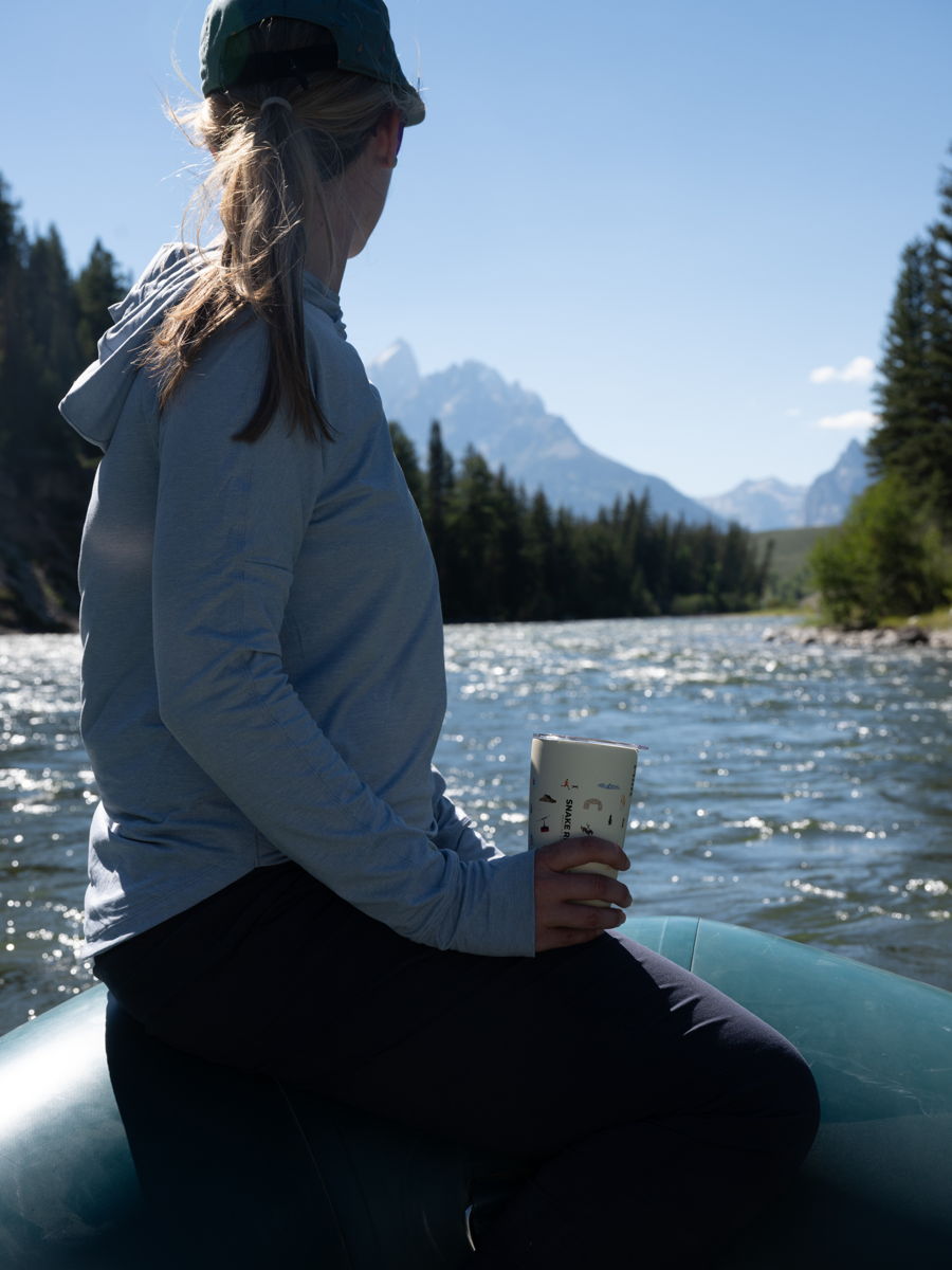Cait Rifkin seated on a raft on the Snake River, coffee cup in hand, with the Tetons ahead